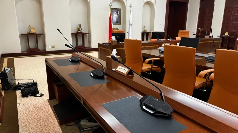 A white conference room with alcoves, dark wood tables and equipped with long-stemmed microphones and discussion devices at each seat.